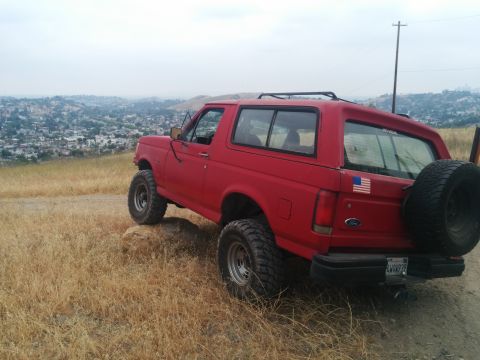 1988 Ford Bronco - A red Ford Bronco is parked on a grassy hill overlooking a city, with its spare tire mounted on the back.