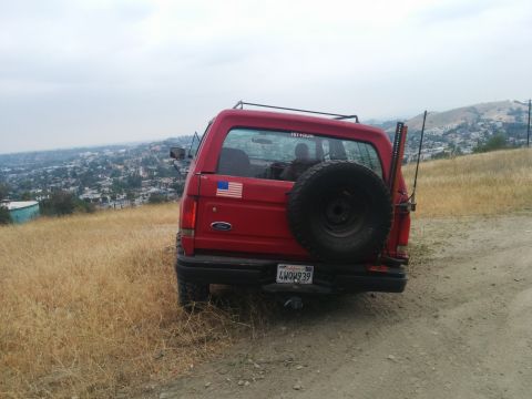 1988 Ford Bronco - Rear view of a red 1988 Ford Bronco parked on a hill overlooking a town, a spare tire on the tailgate and an American flag decal.