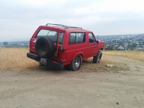 1988 Ford Bronco - Rear view of a red Ford Bronco with a spare tire on the back, parked on a hillside.