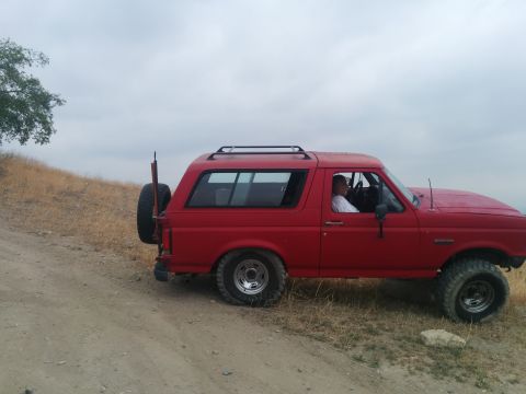 1988 Ford Bronco - A red SUV is parked on a dirt road with a tree and dry grass in the background under a cloudy sky.