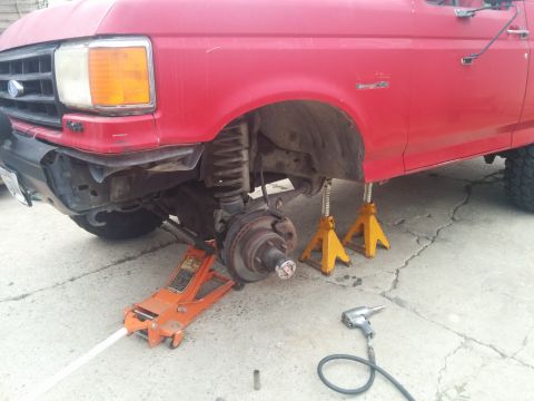 1988 Ford Bronco - Front end of a red truck undergoing maintenance, with the wheel off and supported by jacks.