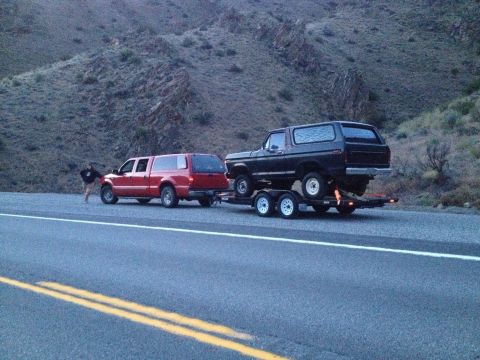 1978 Ford Bronco - A red pickup truck tows a 1978 Ford Bronco on a trailer down a highway.
