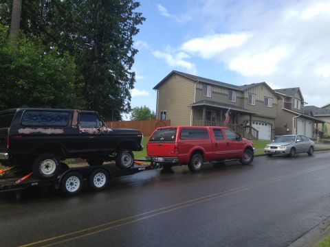 1978 Ford Bronco - A black 1978 Ford Bronco is loaded onto a trailer, towed by a red truck, with a silver sedan parked nearby.