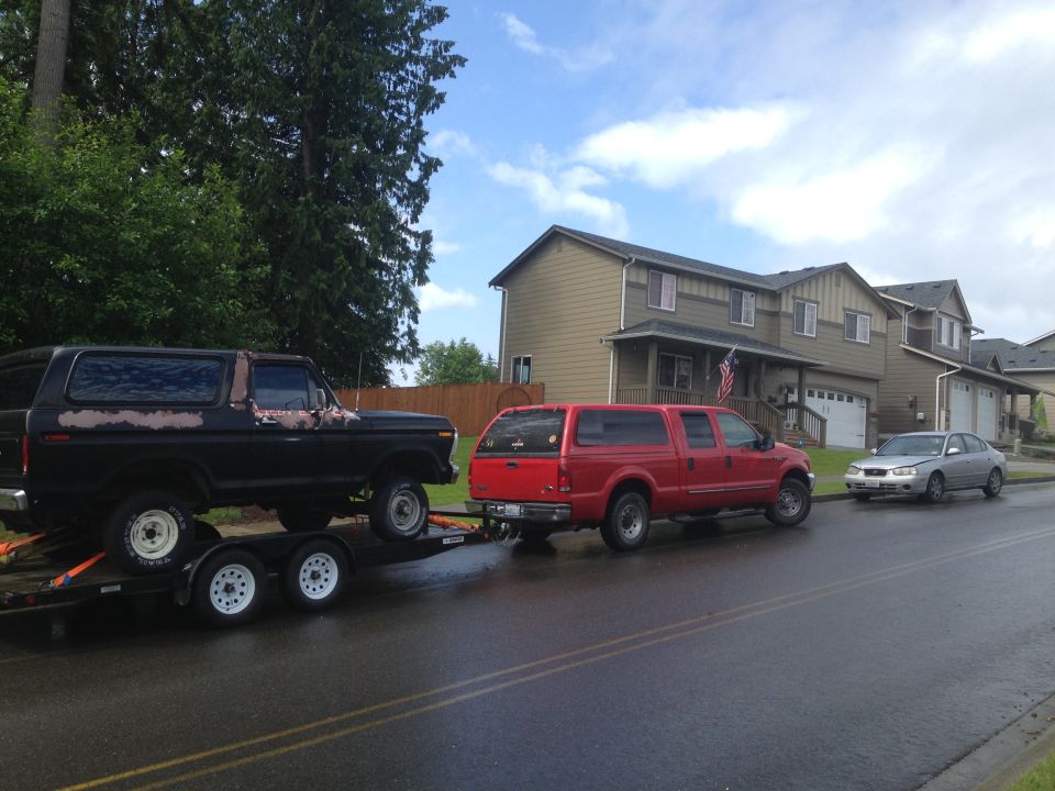 Black 1978 Ford Bronco on a trailer being towed by a red truck, with a silver car parked in front of a house