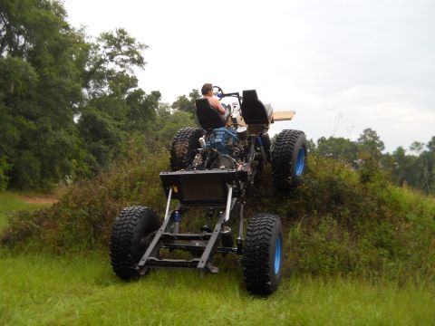 1992 Ford Bronco - A driver navigates an off-road vehicle chassis up a grassy hill, its suspension and large tires.