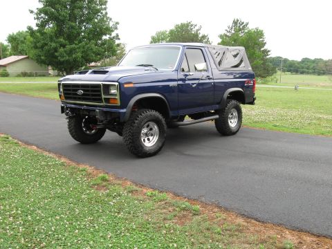 1982 Ford Bronco - This lifted blue Ford Bronco features a soft top and aftermarket wheels, parked on a paved surface with grassy areas on either side.