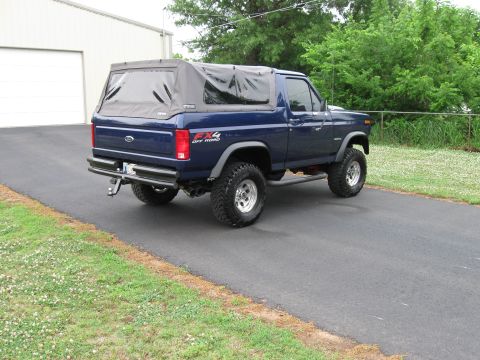 1982 Ford Bronco - This blue Ford Bronco is shown from the rear 3/4 view, parked on a paved driveway next to a grassy area.
