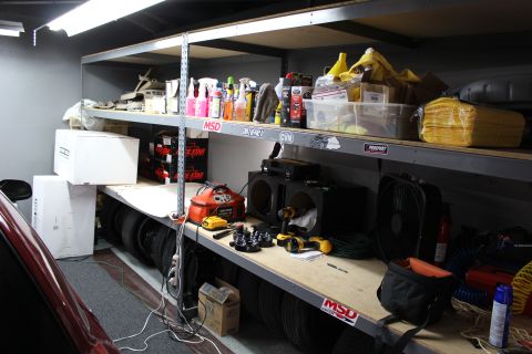 2003 Ford Crown Victoria - cluttered garage shelf holding spray bottles, tools, and stacked boxes above a workbench with a red car at left.