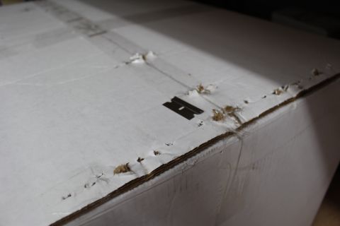 2003 Ford Crown Victoria - Detail of a white shipping box corner showing torn liner, exposed corrugation, and a small black stamped mark.