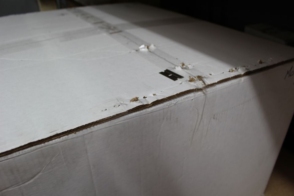 White cardboard box top with seams and a small black stamped marking, frayed edges
