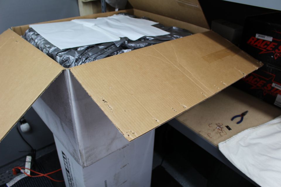 Open cardboard box with white-paper-covered contents in a workshop near red shelving boxes