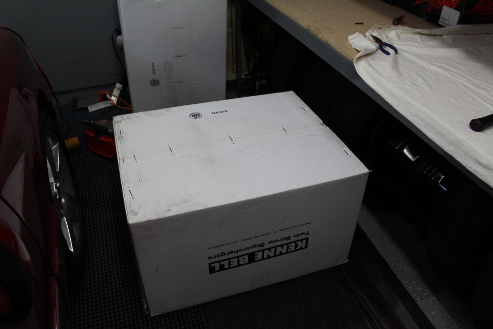 White Kenne Bell supercharger box on shop floor beside a red car and wheel