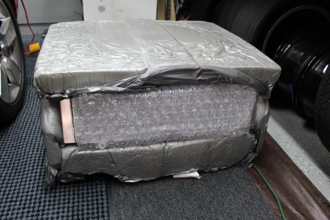 2003 Ford Crown Victoria - bulky item wrapped in silver foil insulation, bubble wrap, and gray tape sits on a shop floor by a wheel.