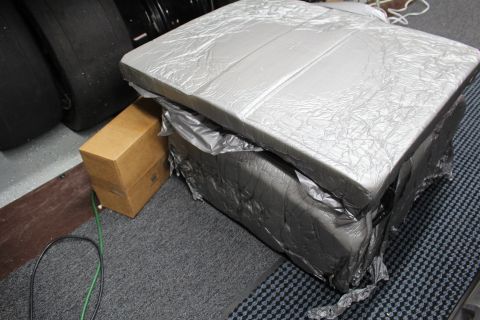 2003 Ford Crown Victoria - foil-topped bundle wrapped in dark plastic sits on the shop floor next to a small brown box and tires.