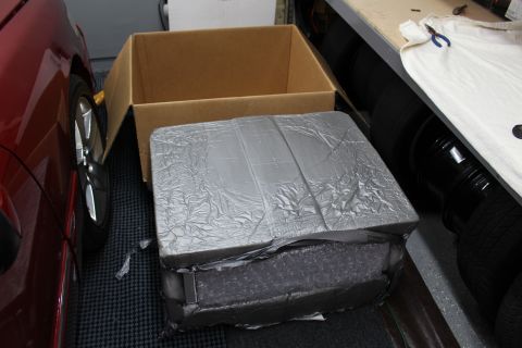 2003 Ford Crown Victoria - foil-and-bubble-wrapped item placed atop an empty open cardboard box next to a red car's front wheel.