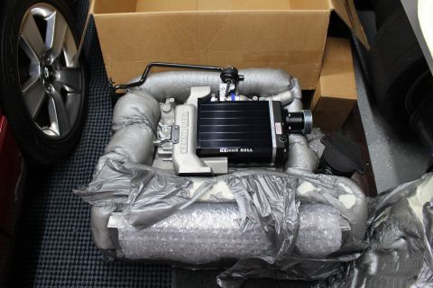 2003 Ford Crown Victoria - black-and-aluminum Kenne Bell supercharger sits nestled in gray foam packaging inside a shipping box beside a five-spoke alloy wheel.