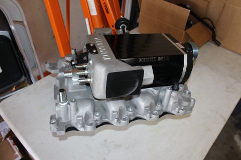 2003 Ford Crown Victoria - End view of a Kenne Bell supercharger showing the black inlet opening and Kenne Bell-branded aluminum manifold on a bench.