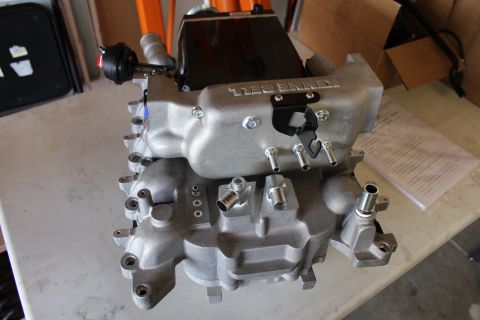 2003 Ford Crown Victoria - bare cast-aluminum supercharger lower intake manifold rests on a shop counter beside paperwork, with mounting bosses and ports visible.