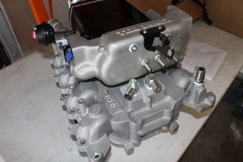 2003 Ford Crown Victoria - unpainted cast-aluminum supercharger manifold base shows protruding nipples, threaded fittings and bolt holes on a workbench.