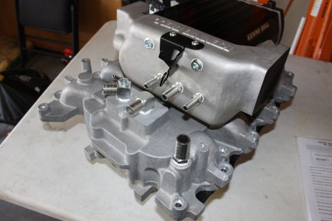 2003 Ford Crown Victoria - cast-aluminum manifold base topped with a black housing labeled Kenne Bell, fitted with studs and ports, sits on a bench.