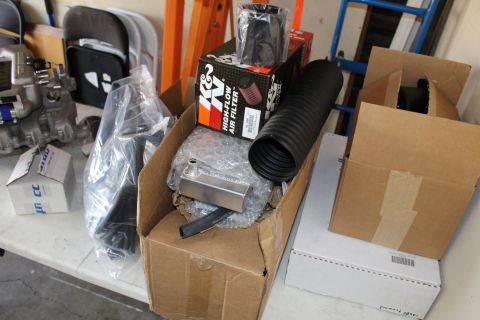2003 Ford Crown Victoria - Boxed supercharger kit parts including a K&N air filter, flex tubing, and an aluminum bracket spread across a garage workbench.