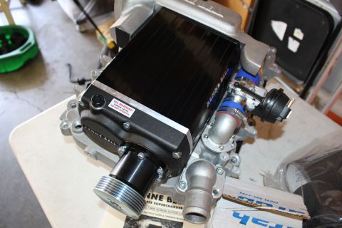 2003 Ford Crown Victoria - black finned Kenne Bell supercharger with warning label and front drive pulley sits on a workbench in a garage.