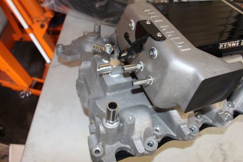 2003 Ford Crown Victoria - cast-aluminum intake manifold rests on a shop bench, fitted with fuel-line fittings and a black throttle-body inlet.