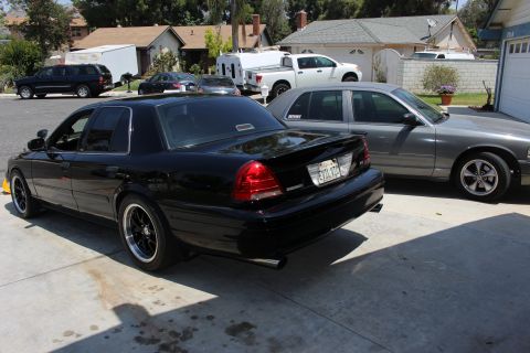 2003 Ford Crown Victoria - black full-size sedan with dark wheels and a deck spoiler sits in a residential driveway beside a silver sedan.