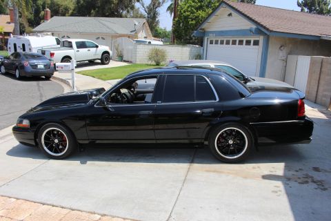 2003 Ford Crown Victoria - Driver-side profile of a black four-door sedan lowered on black wheels, parked in a suburban driveway.