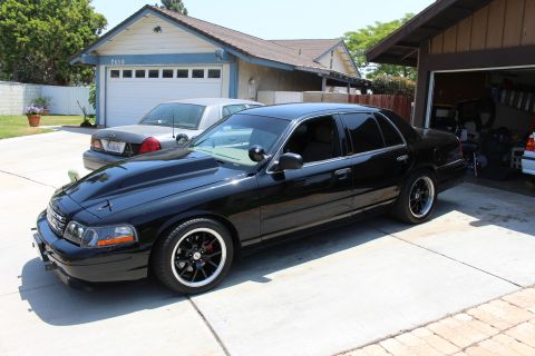 2003 Ford Crown Victoria - black Ford sedan with a raised cowl-style hood and black wheels sits angled in a driveway near a garage.