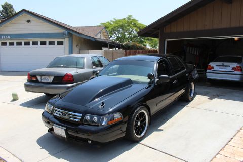 2003 Ford Crown Victoria - black Ford four-door with a scooped cowl hood and black wheels parked in front of a garage next to a gray sedan.