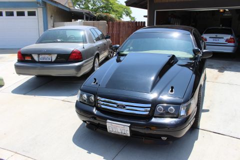 2003 Ford Crown Victoria - black Ford sedan with a cowl hood wears a front plate reading EVIL VIC while parked beside a gray sedan.