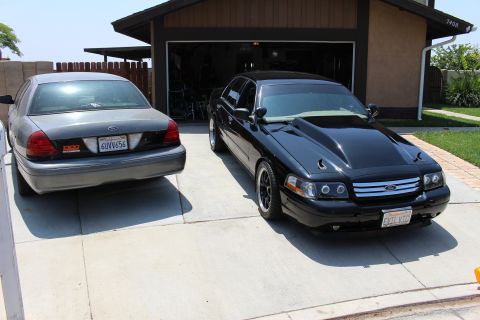 2003 Ford Crown Victoria - black Ford sedan with a raised cowl hood parks beside a gray Ford sedan on a residential driveway.