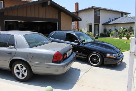 2003 Ford Crown Victoria - gray Ford sedan and a black Ford sedan on black wheels sit side by side in a suburban driveway.