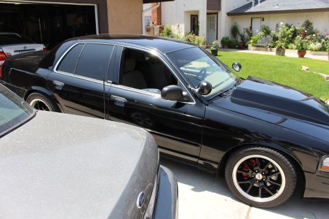 2003 Ford Crown Victoria - Passenger-side view of a black Ford sedan with tinted glass and black wheels, parked next to a gray car.