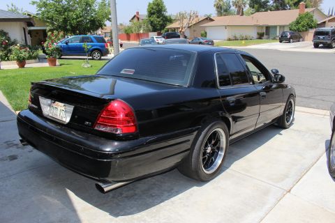 2003 Ford Crown Victoria - black full-size sedan with a deck-lid spoiler and black wheels shown from the rear right in a driveway.