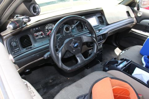 2003 Ford Crown Victoria - modified Ford sedan interior shows a small aftermarket steering wheel, dash-mounted display, and orange-trimmed seat.