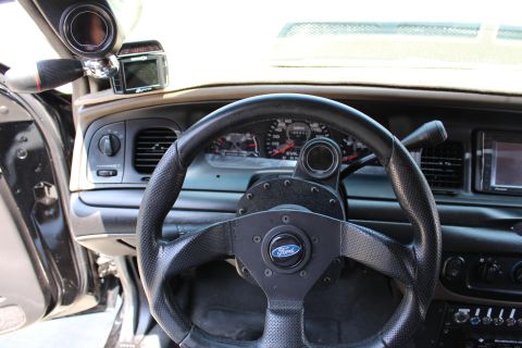 2003 Ford Crown Victoria - Ford-badged aftermarket steering wheel sits before a gauge cluster reading EVIL VIC, with an A-pillar gauge alongside.