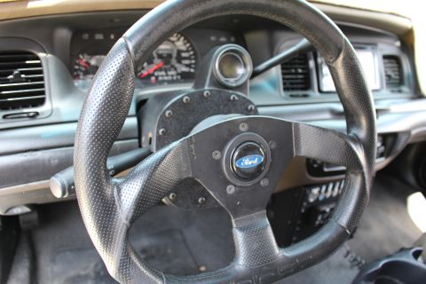 2003 Ford Crown Victoria - Small black perforated aftermarket steering wheel fitted with a Ford center emblem, mounted in front of the gauge cluster.