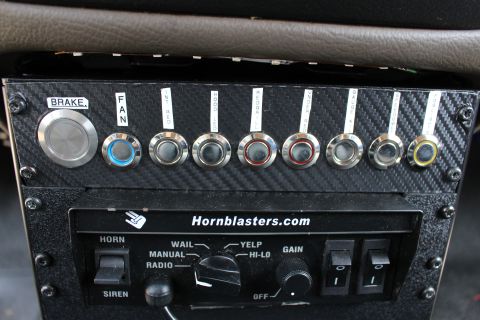 2003 Ford Crown Victoria - Detail of a HornBlasters horn and siren control unit beside labeled chrome-ringed toggle switches on a carbon-fiber panel.
