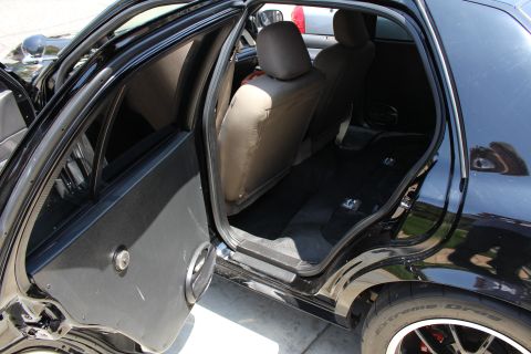 2003 Ford Crown Victoria - Open rear door of a black sedan showing the back seat with tan covers, door panel, and a dark alloy wheel.