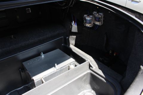 2003 Ford Crown Victoria - Sedan trunk interior showing a metal storage box, clear audio capacitors, and bundled wiring mounted on the carpeted side wall.