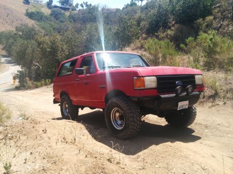 1988 Ford Bronco - A red Ford Bronco is seen from a 3/4 front angle on a dirt road with hills in the background.