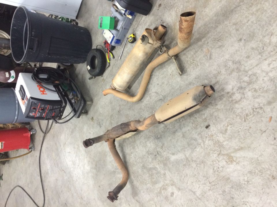 Two rusty exhaust assemblies with mufflers on a garage floor near a Hobart welder and tools
