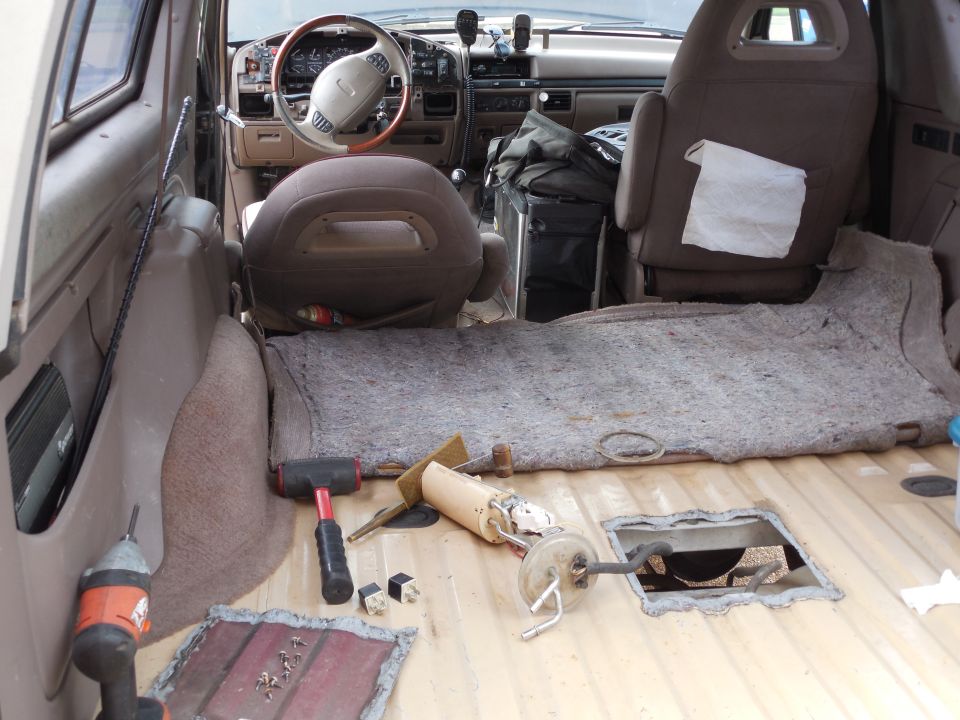 Truck interior with carpet pulled up, floor access holes open, and fuel pump parts and tools on the bare floor.