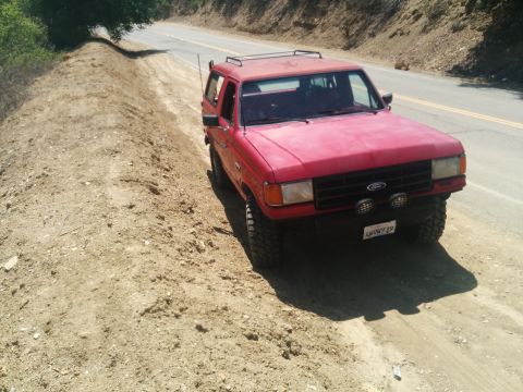 1988 Ford Bronco - This red 1988 Ford Bronco is parked on a dirt road with a tree overhead.