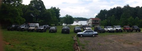 1986 Ford Bronco - A collection of SUVs and a sedan are parked on a grassy area with a lake and house in the background.