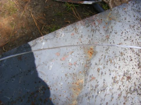 Close-up of a rusty, weathered metal panel with a seam. Some dirt and grass visible in the background. Waterma
