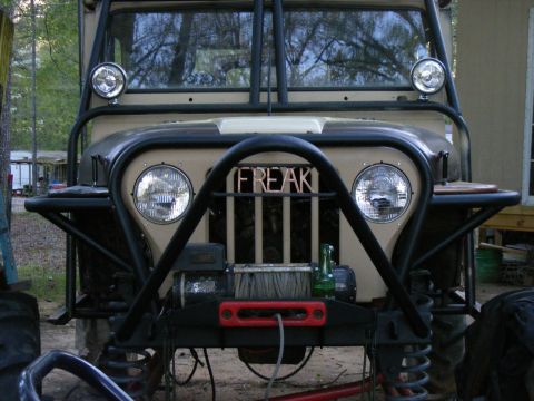 Front view of a tan off-road Jeep with a black roll cage, winch, and 'FREAK' spelled out on the grille