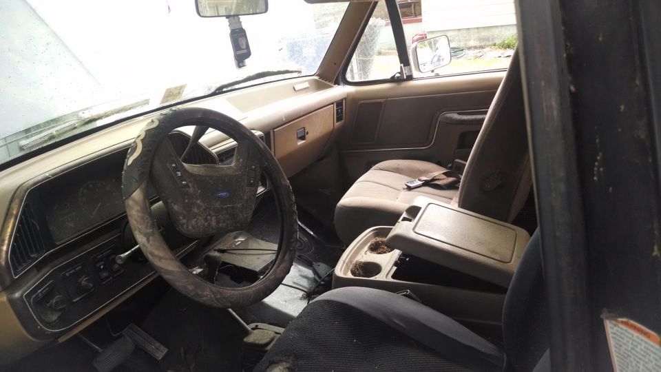 1987 Ford Bronco interior, tan dashboard, dirty steering wheel, open center console with cup holders, worn seats